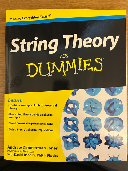 The front cover of the book 'String Theory For Dummies' by Andrew Zimmerman Jones, featuring a black and yellow design with blue abstract illustrations, photographed on a wooden surface.